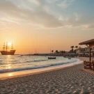 جاهای دیدنی کیش،غروب ساحل مرجان کیش با آب‌های فیروزه‌ای و کشتی یونانی در دوردست، زیر نور نارنجی و صورتی آسمان.